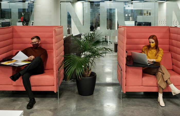 Two people in modern office booths wearing face masks, working on documents and laptop, maintaining focus.