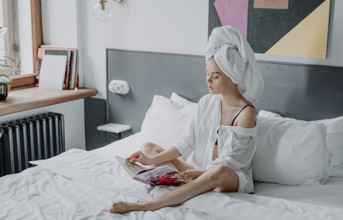 A young woman with washed hair wrapped in a towel, reading a magazine on a comfortable white bed.