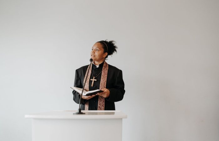 A female pastor delivers a sermon at a church podium, holding a Bible and wearing a cross.